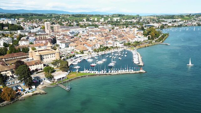 Aerial view of Morges Castle and harbor with boats docked on turquoise waters, contrasting with the red rooftops of the city buildings, Morges, Vaud, Switzerland.