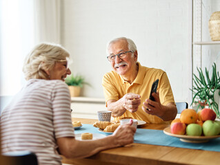 Portrait of an elderly senior couple having breakfast and using a smart phone at home. Happy healthy affectionate senior couple talking and showing smartphone screen to one another and texting and sit