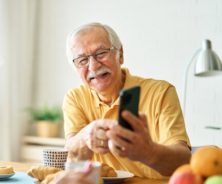 Portrait of an elderly senior couple having breakfast and using a smart phone at home. Happy healthy affectionate senior couple talking and showing smartphone screen to one another and texting and sit - Powered by Adobe