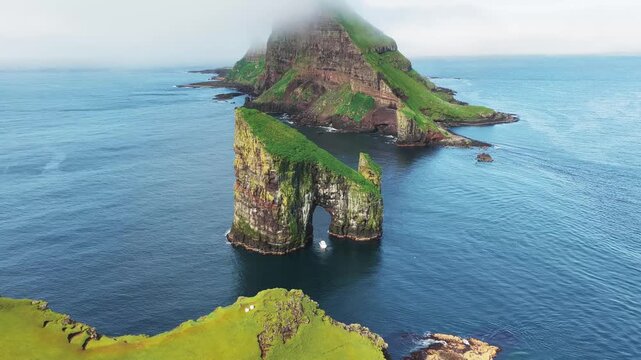 Aerial view of the majestic Drangarnir sea stack archway with vibrant green grass contrasting with the deep blue ocean waters, Drangarnir, Faroe Islands.
