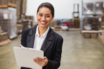 Smiling African American woman in blazer holding clipboard and pen, inspecting stock in warehouse