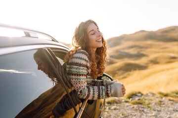 A happy traveler leans out of a car window at sunset. The young woman feels free and enjoys the...