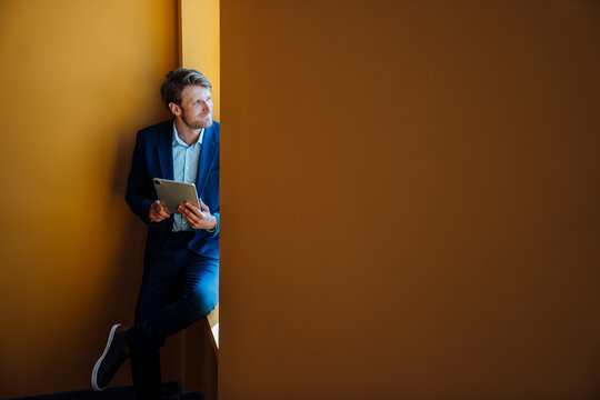 Businessman in formal wear with tablet looking away by window indoors