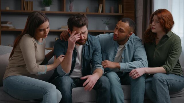 Four diversity friends in living room desperate Caucasian man sharing bad news disappointed problem multiethnic people men women support cuddling attention friendship at home party psychological help