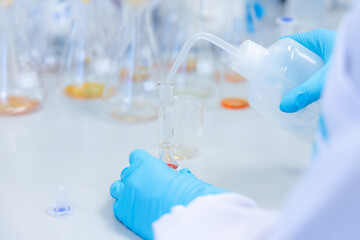 A scientist carefully pours liquid from a laboratory wash bottle into a glass test tube during a chemical experiment. The modern laboratory with various glassware and scientific equipment visible.