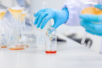 Close-up of hands pouring an orange chemical solution into a beaker, representing chemical analysis, food safety testing, and quality control procedures in a clean and professional lab environment.