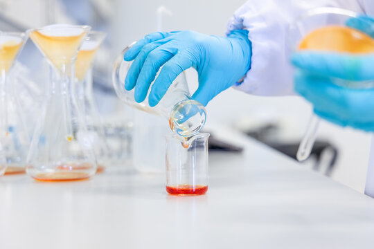 Close-up of hands pouring an orange chemical solution into a beaker, representing chemical analysis, food safety testing, and quality control procedures in a clean and professional lab environment.