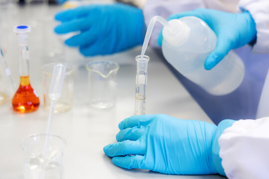 A scientist carefully pours liquid from a laboratory wash bottle into a glass test tube during a chemical experiment. The modern laboratory with various glassware and scientific equipment visible.