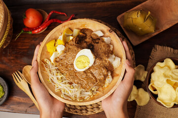 Traditional Indonesian kupat tahu served in a wooden bowl with rice cake, tofu, bean sprouts, and boiled egg topped with rich peanut sauce and crunchy crackers.