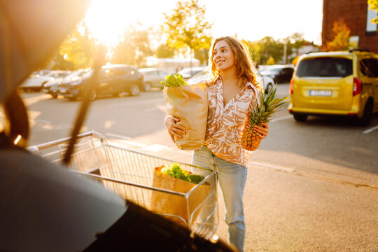 Beautiful woman stands with bag of fresh fruits and vegetables in sunny parking lot near car. A woman with a cart and a paper bag of groceries enjoys the sunset. Concept is thrifty and fresh.