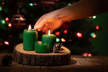A hand uses a match to light green candles on a wooden surface with a Christmas Tree in the background decorated with lights and ornaments. The scene evokes a festive atmosphere.