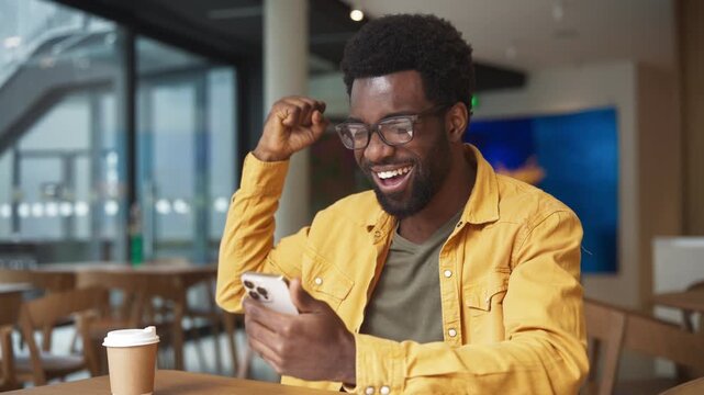 Excited African American man wearing glasses laughing while checking smartphone in cafe. Male smiling with joy looking at phone screen, celebrating success with coffee cup. Victory or good news.