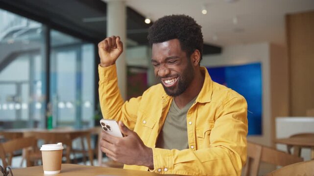 Happy African American man celebrating success looking at smartphone in cafe. Male raising fist and laughing with excitement while checking good news indoors. Joyful moment at coffee shop. Victory.