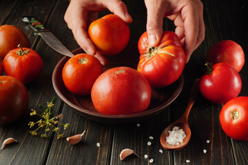 A person sorts ripe, red tomatoes from a bowl onto a wooden table. Garlic and salt are nearby,...