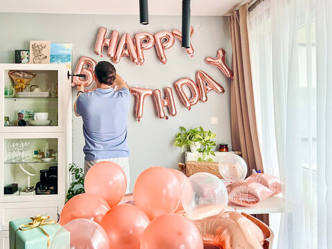 Father decorating living room with balloons and happy birthday banner, preparing space for teenage daughter’s birthday celebration. Cozy family home atmosphere, authentic candid moment.