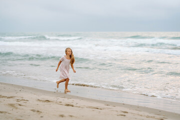 A cute girl in a dress is having fun on the seashore. The girl enjoys the seascape, runs along the seashore. The concept of childhood, fun. Lifestyle.