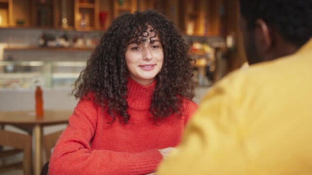 Caucasian woman with curly hair leaning on hand while talking indoors at cafe. Female engaged in pleasant conversation with African American man, sitting at wooden table in modern coffee shop.
