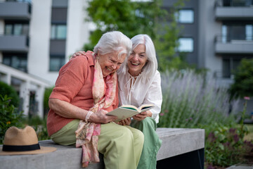 Elderly mother sitting on bench with adult daughter, reading together.