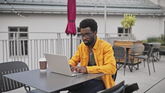 Handsome African American man with beard wearing glasses working on laptop outdoors at table. Male concentrating on computer with coffee cup beside, enjoying remote work outside.