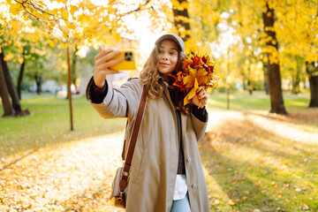 A young woman in an autumn raincoat takes a selfie on her phone in a sunny park. Autumn mood. A smiling woman with a bouquet of colorful leaves has fun and blogs on her phone on an autumn day.