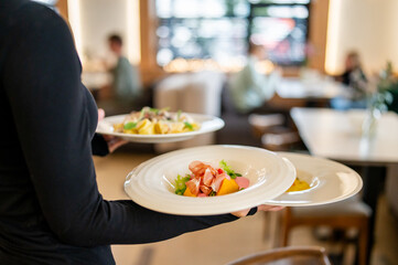 Restaurant server in black uniform carrying three plated meals, including salad, pasta, and polenta, in a modern dining setting with blurred guests and wooden furniture
