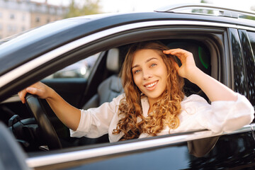 Portrait of smiling woman sitting behind the wheel of car looking out the window. Woman driver in modern car enjoying sunny day. Transport, taxi concept. Lifestyle.