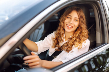 Portrait of smiling woman sitting behind the wheel of car looking out the window. Woman driver in modern car enjoying sunny day. Transport, taxi concept. Lifestyle.