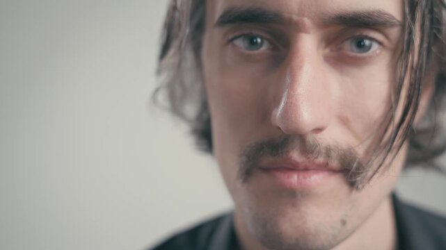 A light smile and skeptical squint highlight a young handsome man showcasing a range of emotions through his eyes and mustache in a neutral background.