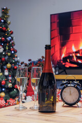 Two champagne glasses, a bottle and clock on a festive table with Christmas decorations and fireplace light, symbolizing New Year celebration