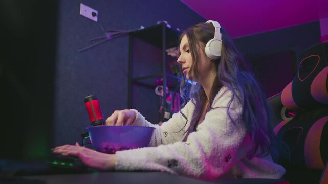 Young woman wearing headphones types on keyboard while eating popcorn from bowl, wipes hand on fluffy cardigan, then resumes typing in colorful neon lit workspace