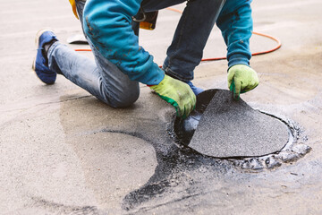 Roofer repairing a damaged section of a roof. Worker kneels on a wet, flat rooftop to apply a...