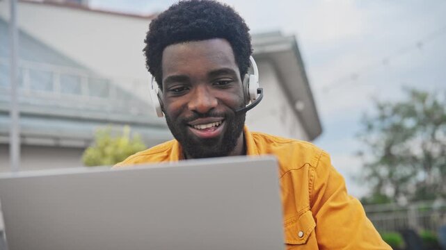 Handsome African American man with beard wearing headset talking while working on laptop outdoors. Male communicating online, sitting near modern building and enjoying remote work outside.