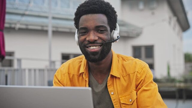 Portrait of handsome African American man with beard wearing headset smiling while working on laptop outdoors. Male enjoying remote communication, sitting near modern building outside.
