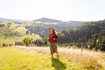Smiling woman standing on a lush green field surrounded by mountains. Young female traveler with a backpack enjoying mountain scenery and walking in the fresh air. Relaxation concept.