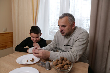 Father and son talk at the table while cleaning walnuts, the child smiles as the man holds a cleaned piece.