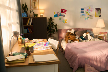No people view of pictures at desk in cozy bedroom, colorful artwork and stuffed animals on bed, sunlight streaming through window