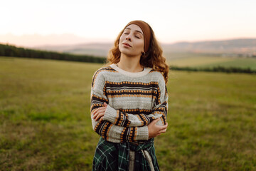 Young woman in a sweater standing on a green hill in the rays of sunset. Traveler enjoying nature and mountain landscape. Nature, recreation concept.