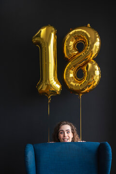 Surprised young woman hiding behind chair with number 18 balloon on black backdrop