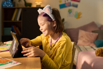 Caucasian girl child smiling while using digital tablet at desk, wearing cat ear headband and necklace, engaging in creative activity in cozy home environment with colorful decorations
