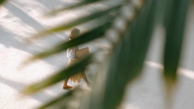 Tropical leaves partially cover a barefoot female digital nomad walking on white sand while carrying a laptop, suggesting a work and travel lifestyle