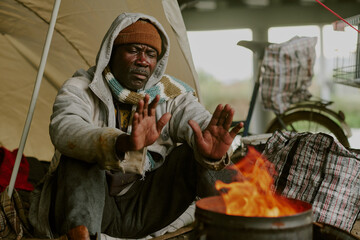 Middle aged Black man sitting outdoors under makeshift shelter warming hands over open fire, wearing layered clothing and knit hat, shopping cart and bags visible in background