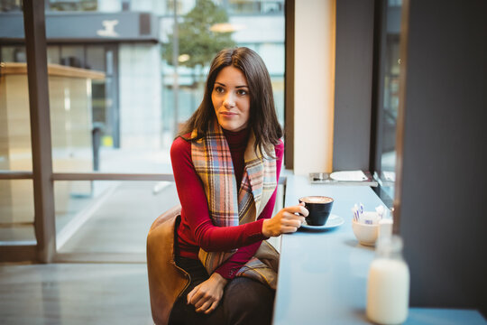 Woman in mid-20s sitting at cafe counter holding coffee mug, using smartphone by window