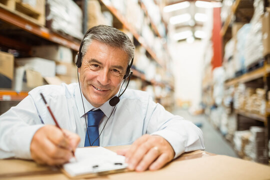 Senior man wearing tie writing on clipboard in warehouse aisle with shelving and headset microphone - Powered by Adobe