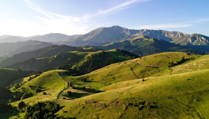 Obraz premium Aerial view of rolling green hills with a winding road leading towards a mountain range under a clear, sunny sky