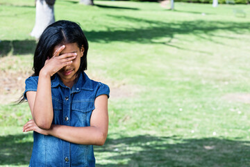 Preteen girl standing near tree trunk on sunlit lawn in park covering face and crossing arm