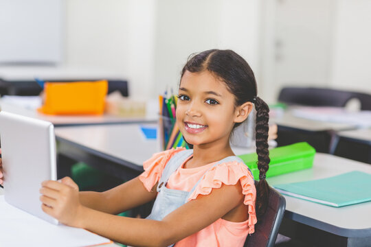 African American schoolgirl smiling, holding tablet at classroom desk with pencils and green binder
