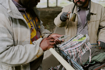 Middle aged Black man holding mobile phone next to middle aged Caucasian man drinking from metal cup, both standing by shopping cart filled with personal belongings outdoors