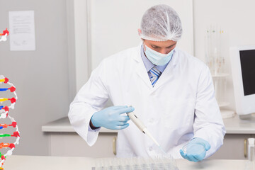 Scientist pipetting samples into multi-well plate near DNA model and computer monitor at lab bench