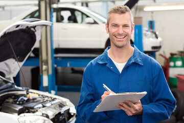 Male mechanic smiling, holding clipboard and pen in workshop beside open engine bay