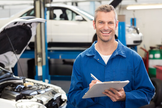 Male mechanic smiling, holding clipboard and pen in workshop beside open engine bay
